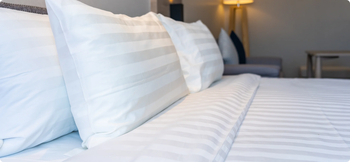 A close-up of a neatly made bed with white and light gray striped bedding, including pillows and a duvet, in a softly lit bedroom setting.