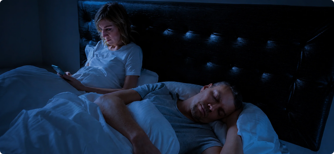 A man sleeps soundly in bed while a woman sits up beside him, holding a phone in a dimly lit bedroom with a dark tufted headboard.