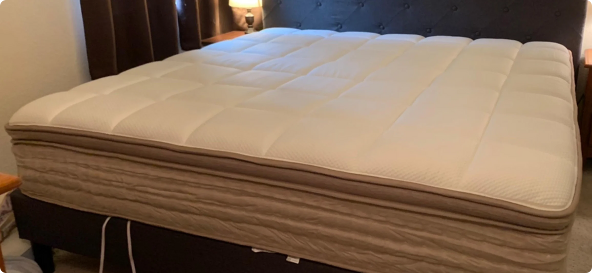 A close-up view of a mattress topper or mattress surface on a bed frame in a bedroom setting, with a dark upholstered headboard and bedside lamp visible in the background.