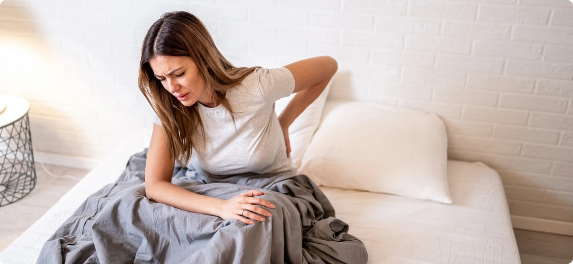 A woman sitting on a bed in a bright bedroom, holding her lower back in discomfort. She is wearing a light gray t-shirt and a white pillow is behind her.