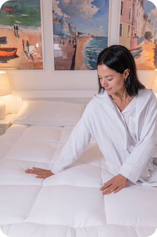 A woman in a white robe sits on a white quilted bed, touching the mattress. The bedroom has a white headboard, bedside lamps, and three colorful framed paintings on the wall depicting coastal scenes.