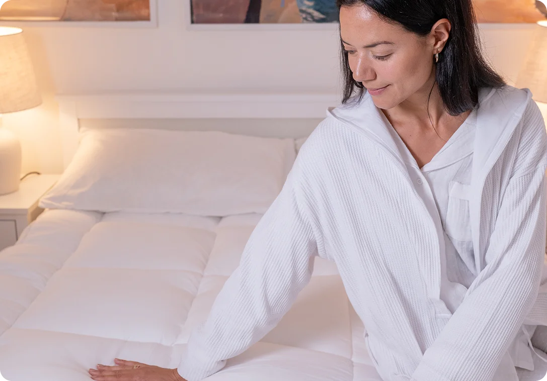 A woman wearing a white ribbed hooded robe sits on a white bed in a softly lit bedroom, touching the quilted bedding.